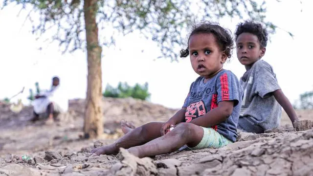 Two young Sudanese children sitting on dry, cracked ground under a tree, with a seated man in the background, file photo