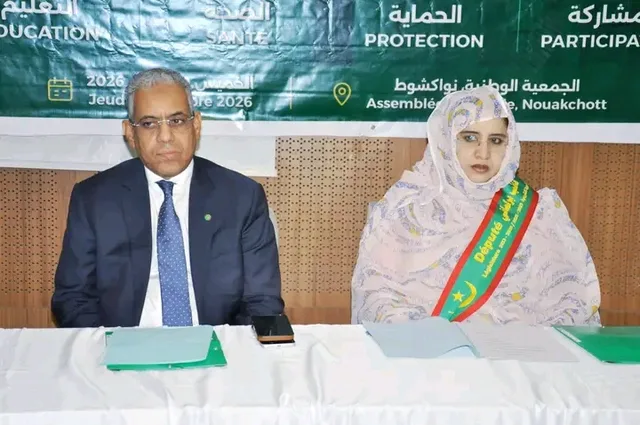 An official and an MP wearing a sash in front of the meeting banner