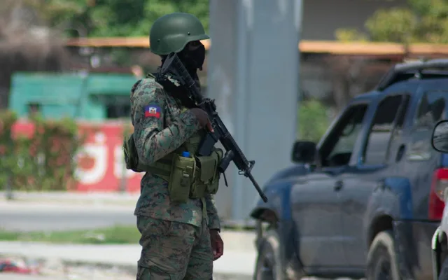 A masked security officer in fatigues and helmet holding a rifle, a Haitian flag patch on his shoulder, near an SUV, file photo