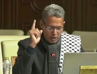 An MP wearing glasses speaks into a microphone in a parliamentary chamber