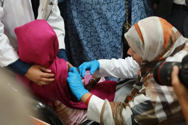 A gloved health worker vaccinates a child held by an adult