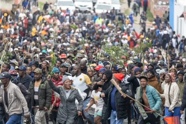 A large crowd of protesters, some carrying sticks and branches, marches down a street in South Africa during an anti-immigrant protest