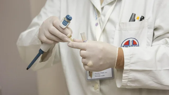 Gloved hands holding a pipette and a sample tube in front of a white coat, illustrative image