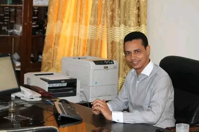 A smiling man seated at a desk with a computer and a printer
