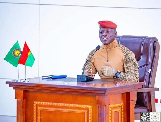 Ibrahim Traore in military uniform and red beret, speaking at a desk in front of a microphone beside the flags of Burkina Faso and the AES, file photo
