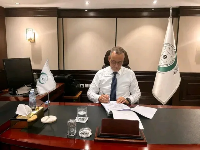 The Secretary-General of the Organisation of Islamic Cooperation signs documents at his desk beside the organisation's flags