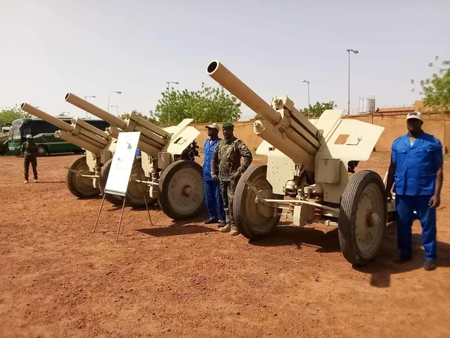 Three field guns on red laterite ground next to a soldier and workers in blue overalls, file photo