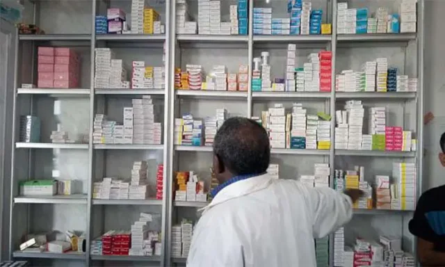 A pharmacist in a white coat, seen from behind, takes a box from shelves stocked with medicines, file illustration