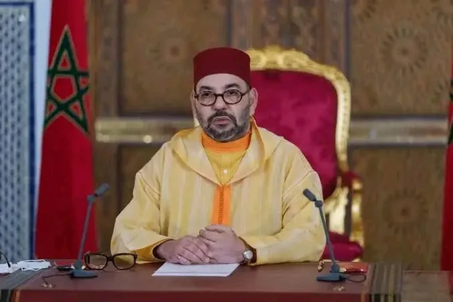 King Mohammed VI seated at a desk in front of Moroccan flags