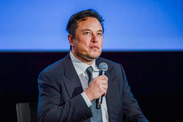 Elon Musk speaking into a microphone against a blue backdrop