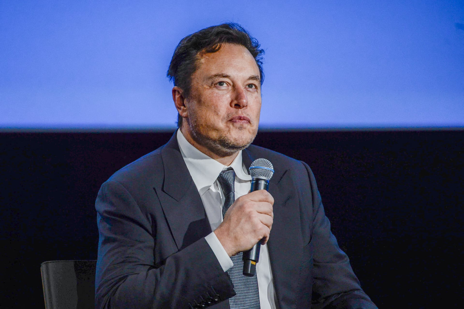 Elon Musk speaking into a microphone against a blue backdrop