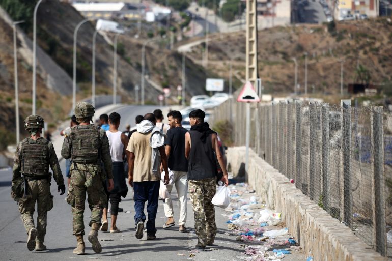 A group of young men walk along a border fence escorted by two soldiers, file photo