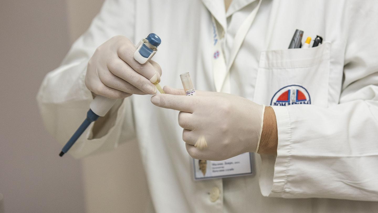 Gloved hands holding a pipette and a sample tube in front of a white coat, illustrative image