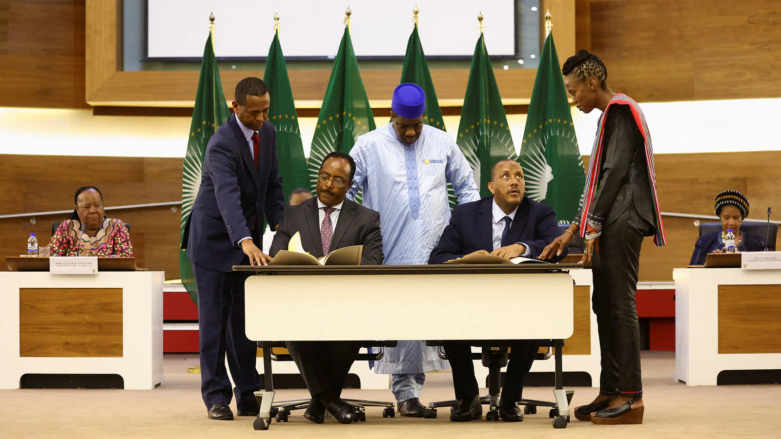 Two seated representatives signing documents at a white table, flanked by mediators in front of green African Union flags, file photo