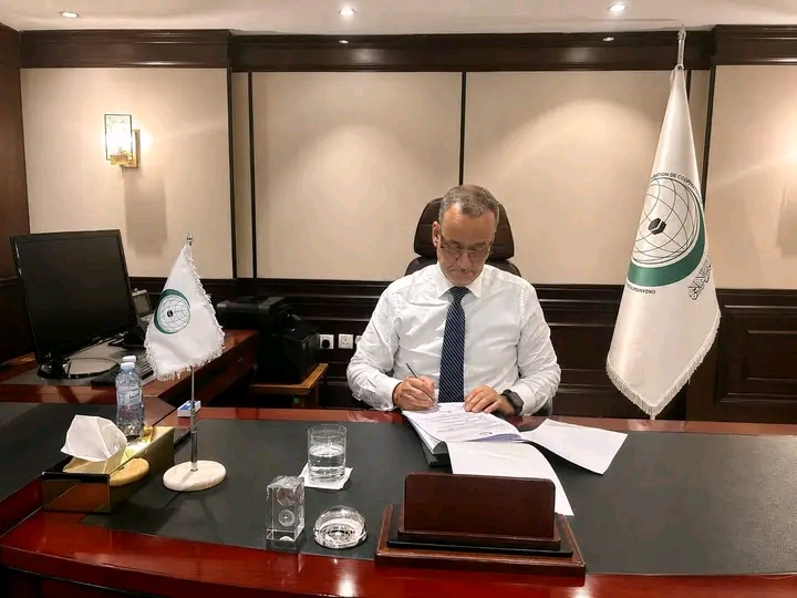 The Secretary-General of the Organisation of Islamic Cooperation signs documents at his desk beside the organisation's flags