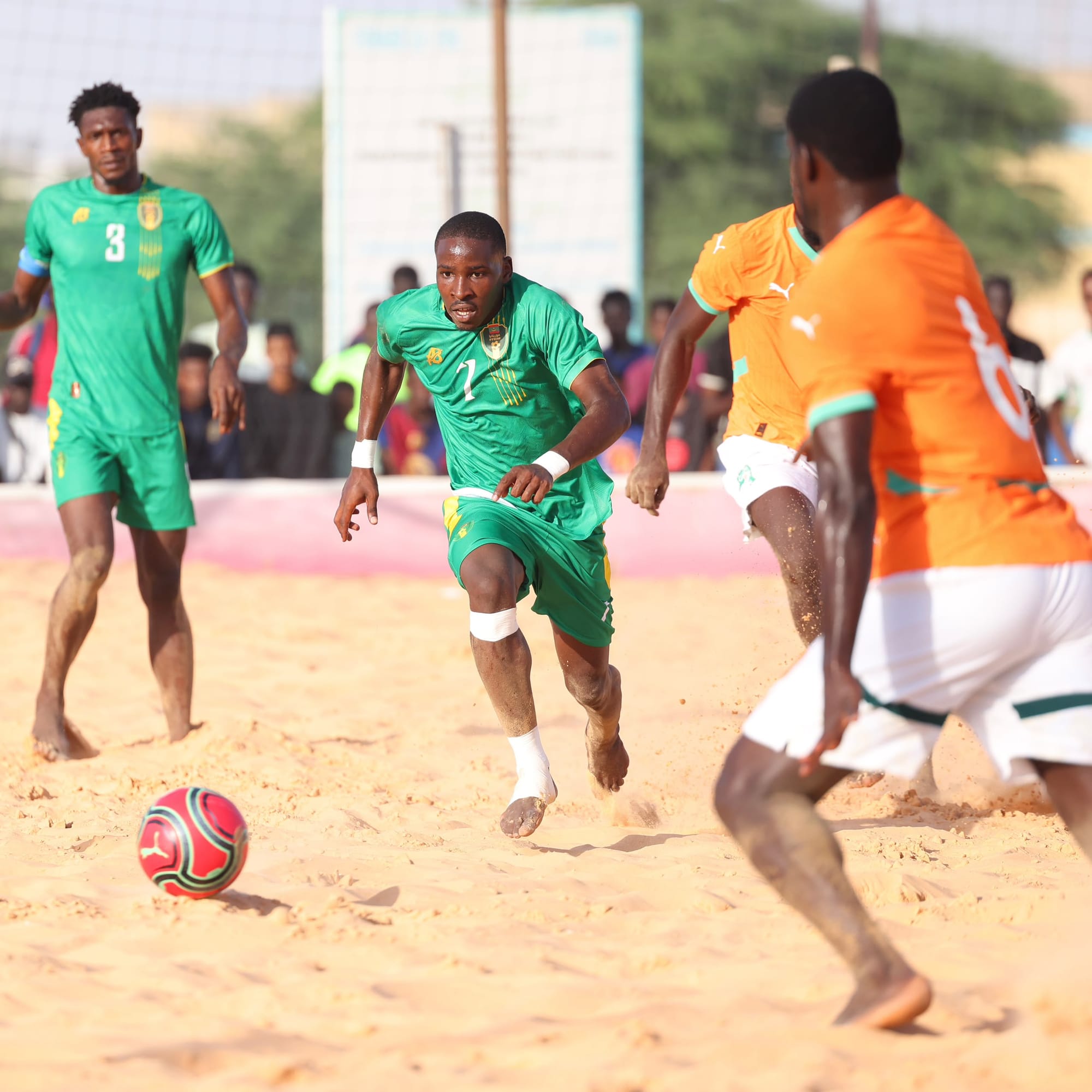 A Mauritanian player in green runs with the ball against Ivorian players on the sand