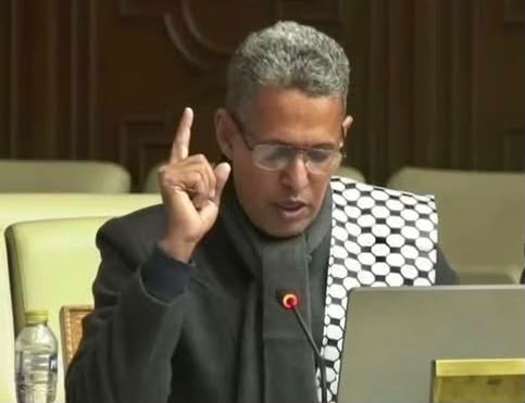 An MP wearing glasses speaks into a microphone in a parliamentary chamber