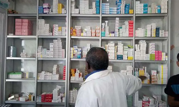 A pharmacist in a white coat, seen from behind, takes a box from shelves stocked with medicines, file illustration