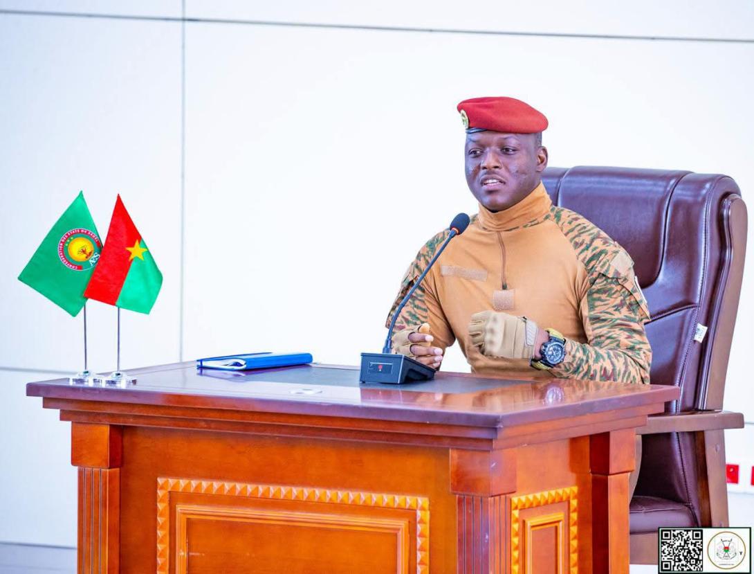 Ibrahim Traore in military uniform and red beret, speaking at a desk in front of a microphone beside the flags of Burkina Faso and the AES, file photo